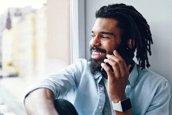 Šťastný Mladý Afričan Mluví Smartphone Usmívá Zatímco Sedí Parapetu Uvnitř — Stock fotografie