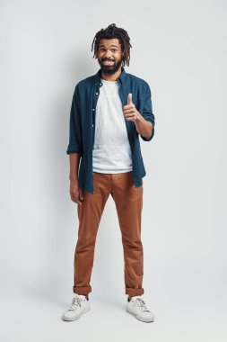 Gri arka planda dikilirken gülümseyen ve baş parmağını gösteren gündelik giysili yakışıklı Afrikalı bir adam.