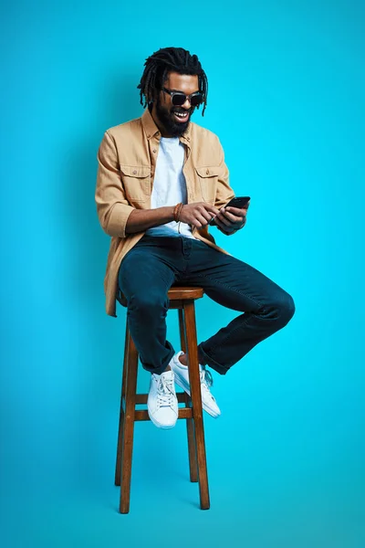 青を背景に座っている間にスマートフォンを使用してカジュアルな服装で魅力的な若いアフリカ人男性の完全な長さ — ストック写真