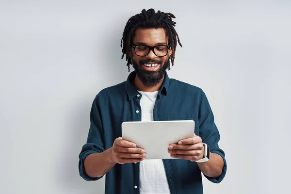 身穿眼镜的忙碌的非洲年轻人一边站在灰色背景下 一边用数码平板电脑微笑 — 图库照片