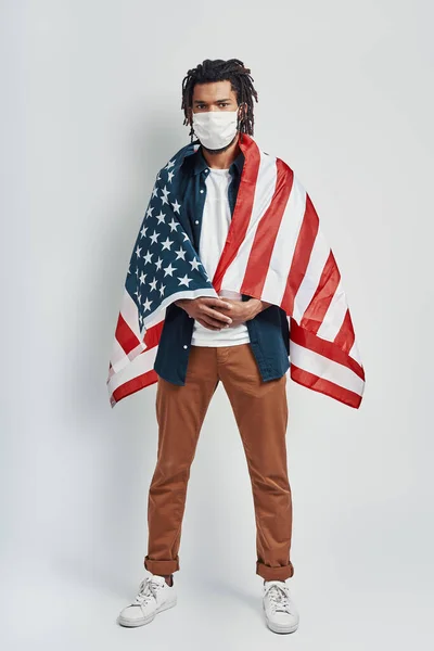 ハンサムな若いアフリカ人男性の完全な長さは 医療面マスクを身に着けているアメリカのフラグで覆われ 灰色の背景に立っている間カメラを見て — ストック写真