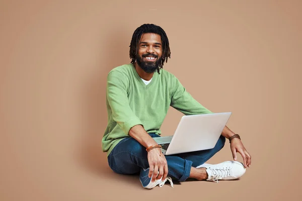年轻英俊的非洲男人 穿着时髦的休闲装 坐在褐色的背景下 一边笑一边用笔记本电脑 — 图库照片