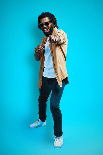 Full length of handsome young African man in casual wear looking at camera and pointing you while standing against blue background             