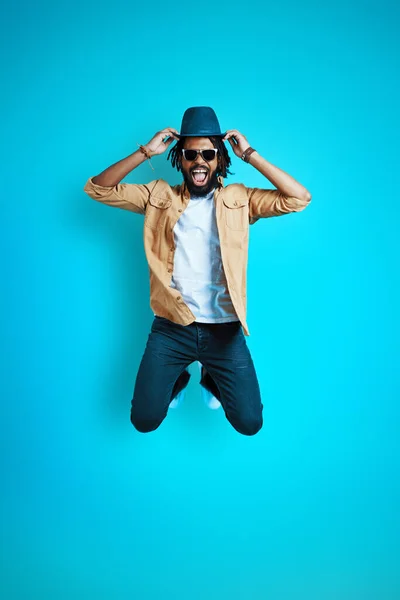 青い背景にホバリングしながら帽子と笑顔を調整カジュアルウェアで遊び心のある若いアフリカの男の完全な長さ — ストック写真