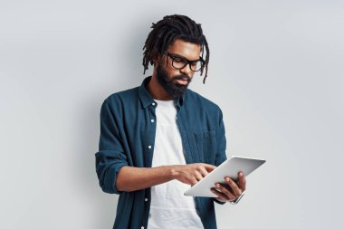 Gri arka planda dikilirken dijital tablet kullanan düşünceli Afrikalı genç bir adam.