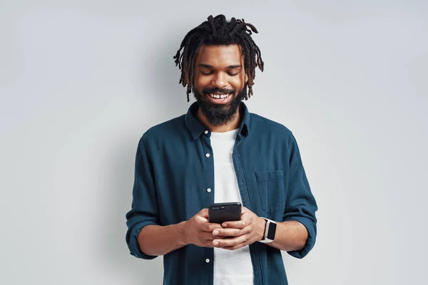 灰色の背景に立っている間に笑顔とスマートフォンを使用して魅力的な若いアフリカ人男性 — ストック写真
