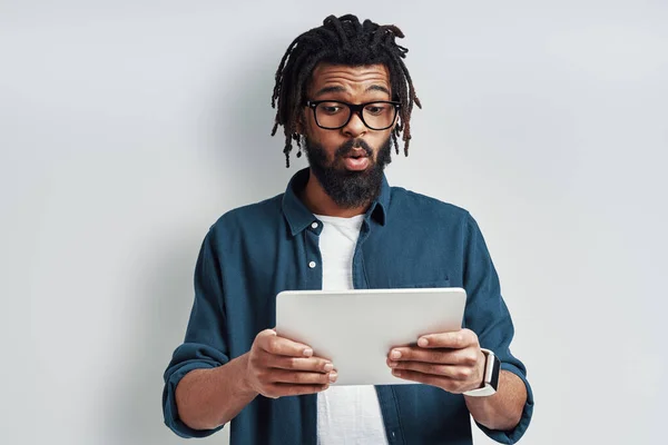 令人惊讶的是 身穿眼镜的年轻非洲男子站在灰色背景下 用数码平板电脑做了个鬼脸 — 图库照片