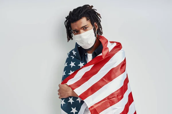 自信を持って若いアフリカ人男性カバーアメリカの国旗を身に着けています医療面マスクとグレーの背景に立っている間カメラを見て — ストック写真