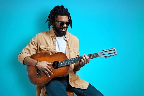 魅力的な若いですアフリカ人男性でカジュアルウェア演奏ギターと笑顔ながら座っている間に青の背景 — ストック写真