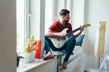Pencere pervazında otururken gitar çalan sıradan giysili yakışıklı bir genç.                