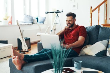 Laptop kullanan ve kanepede otururken gülümseyen rahat giyimli yakışıklı bir genç.    