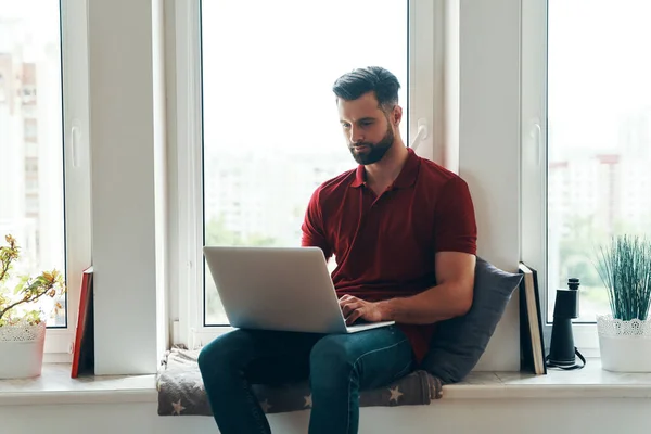 Concentrated young man in casual clothing using laptop while sitting on the window sill