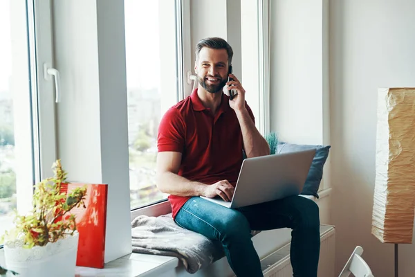 Handsome young man in casual clothing using laptop and talking on the phone while sitting on the window sill