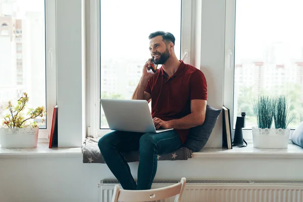 Jovem Feliz Roupas Casuais Usando Laptop Falando Telefone Enquanto Sentado — Fotografia de Stock