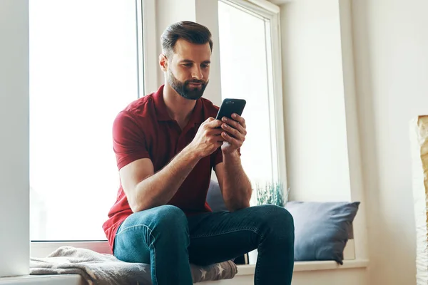 Bonito Jovem Roupas Casuais Usando Telefone Inteligente Sorrindo Enquanto Sentado — Fotografia de Stock