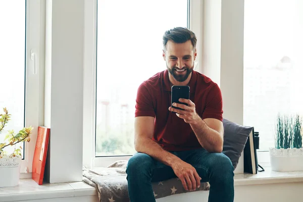 Pemuda Tampan Berpakaian Santai Menggunakan Smartphone Dan Tersenyum Sambil Duduk — Stok Foto