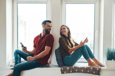 Modern genç çift gündelik kıyafetleriyle birbirine bağlanıyor ve pencerenin pervazında otururken gülümsüyor.