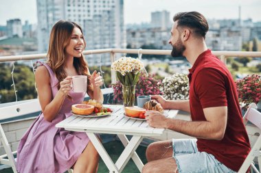 Happy young couple in casual clothing enjoying dinner and smiling while sitting on the rooftop patio outdoors clipart