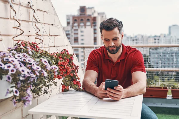 Jovem Bonito Roupas Casuais Usando Smartphone Enquanto Sentado Pátio Telhado — Fotografia de Stock