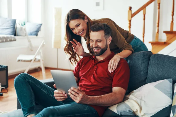 Beautiful young couple in casual clothing talking with someone using digital tablet while resting on the sofa indoors