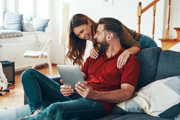 Beautiful young couple in casual clothing talking with someone using digital tablet while resting on the sofa indoors