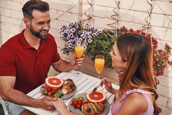 Loving young couple in casual clothing having breakfast and smiling while sitting on the rooftop patio outdoors