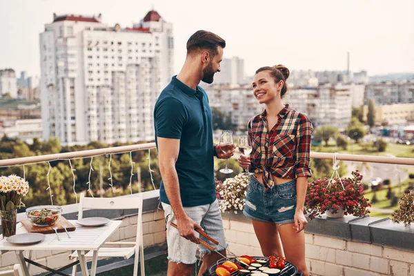 Beautiful young couple in casual clothing preparing barbecue and smiling while standing on the rooftop patio outdoors