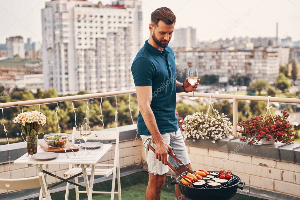 Handsome young man in casual clothing preparing barbecue while standing on the rooftop patio               