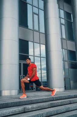 Spor kıyafetleri içinde yakışıklı bir genç adam dışarıda egzersiz yaparken hamlesini yapıyor ve esniyor.