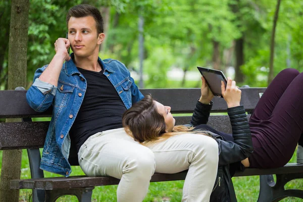 Young men talking on the phone and girlfriend using digital tablet while lying on a bench in park