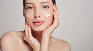 Mavi gözlü ve kahverengi saçlı güzel bir kız yüzüne dokunuyor. Yanaklarına krema sür. Kozmetik ve Spa. Evde bakım.