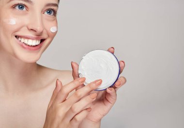 Mavi gözlü genç bir kız gülümsüyor, yana bakıyor ve kremaya parmağıyla dokunuyor. Spa bakım ve güzellik.