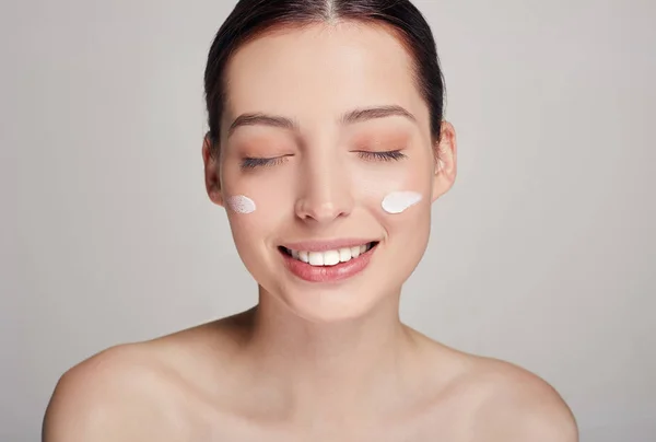 Sonriente joven hermosa chica con el pelo castaño y la piel limpia con los ojos cerrados y crema en las mejillas. Expresiones faciales . —  Fotos de Stock