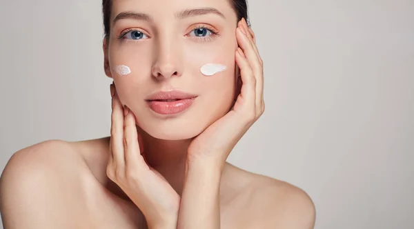 Joven hermosa chica con ojos azules y cabello castaño toca su cara. Crema en las mejillas. Cosmetología y SPA. Atención domiciliaria . —  Fotos de Stock