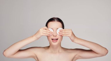 Yakın plan güzel, şaşkın bir kız. Her iki elinde pamuk pedlerle gözlerinin yanında. Arka planda şaşırmış bir ifade var.