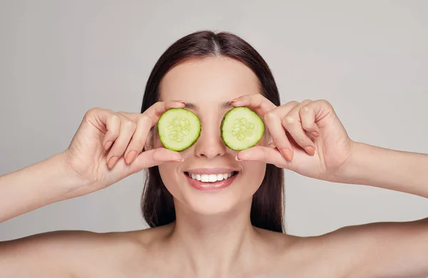 Senhora atraente com cabelo escuro com lábios e um humor brincalhão. A menina fez copos de partes de pepino e sorrindo com dentes brancos. Rejuvenescimento, spa, cosméticos. Espaço de cópia — Fotografia de Stock