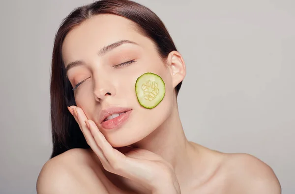 Bela senhorita sedutora com cabelo castanho com lábios cheios e calma completa toca a mão de seu rosto. A menina tem uma fatia fina de pepino na bochecha. Rejuvenescimento, spa, cosméticos . — Fotografia de Stock