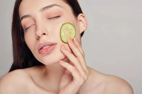 Jovem sedutora bonita com cabelo castanho com lábios cheios e calma total. A menina tem toques pela mão esquerda um fino pepino fresco na bochecha. Rejuvenescimento, spa, cosméticos . — Fotografia de Stock