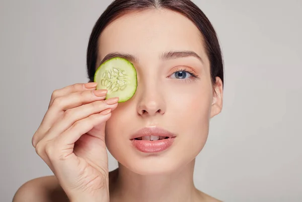 Menina bonita com cabelo castanho com lábios cheios e calma total. A menina segura em sua mão uma fatia de pepino perto de seu olho. Procedimentos de rejuvenescimento. Spa . — Fotografia de Stock