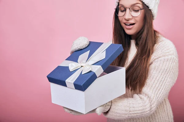 Una joven pensativa se levanta sobre un fondo rosa con gafas, manoplas de color leche y un sombrero, sosteniendo un regalo, que ella abrió y se sorprendió. Preparación para las vacaciones, haciendo bien, Navidad . —  Fotos de Stock