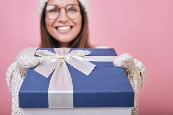 Uma menina feliz alegre está com uma parede traseira rosa em óculos, luvas de leite e chapéu, alcança com um presente para você. Pegue um presente e dê-o a amigos ou parentes. Fixe para dar alegria. Sorrindo. — Fotografia de Stock