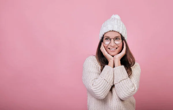 Glada leende flicka med ett utmärkt ansikte kostnader på en rosa bakgrund. damen bär varm stickad tröja och en mjölkfärgad hatt, även i glasögon. Varma kläder under kalla årstider. Mysighet, komfort. — Stockfoto