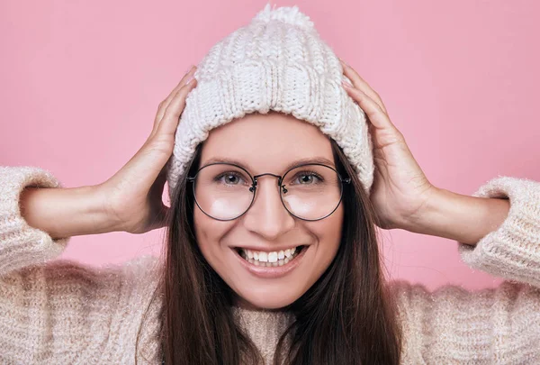 Cheerful smiling girl with excellent face costs on a pink background. lady is wearing a warm knitted sweater and milk-colored hat, also in transparent glasses. Playful mischievous mood, joy of life. — Stock Photo, Image