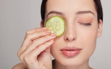 Kahverengi saçlı, dolgun dudaklı ve tamamen sakin genç bir bayan. Kız elinde gözünün yanında bir parça salatalık tutuyor. Gözü kapalı..