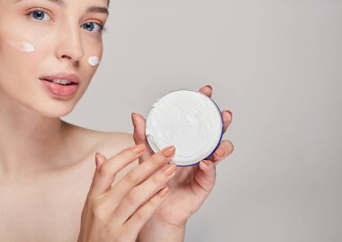 Mavi gözlü genç bir kız, kameraya bakıyor ve kremaya parmağıyla dokunuyor. Evde bakım.