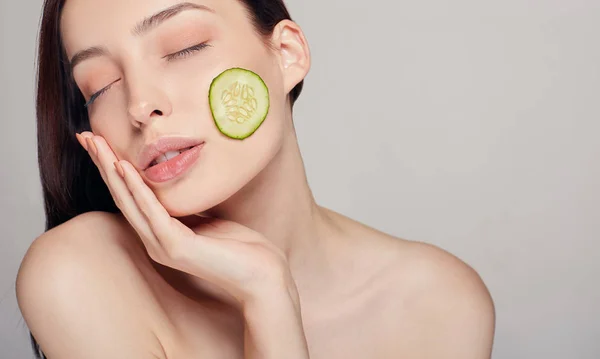 Jovem e sedutora senhora bonita com cabelo castanho com lábios cheios e calma total toca a mão do rosto. A menina tem uma fatia fina de pepino na bochecha. Rejuvenescimento, spa, cosméticos . — Fotografia de Stock