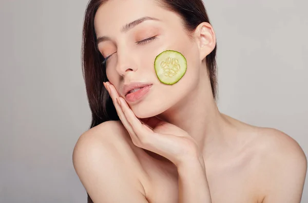 Menina bonita com cabelo castanho com lábios cheios e calma completa toca a mão de seu rosto. As mulheres têm uma fatia fina de pepino na bochecha. Rejuvenescimento, spa, cosméticos . — Fotografia de Stock