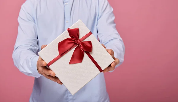 繊細なブルーのシャツを着た男は、ピンクの背景に白い弓で白い蓋をした白い箱に贈り物を持っています。サプライズ、休日. — ストック写真