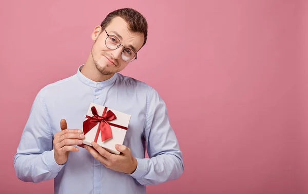 Um homem surpreso de camisa com óculos segura um presente em uma caixa com tampa e arco vermelho em suas mãos, olhando para o presente. Férias, regozijo. Sorrindo, felicidade. Dá-me um presente. O tipo baixou a cabeça. . — Fotografia de Stock