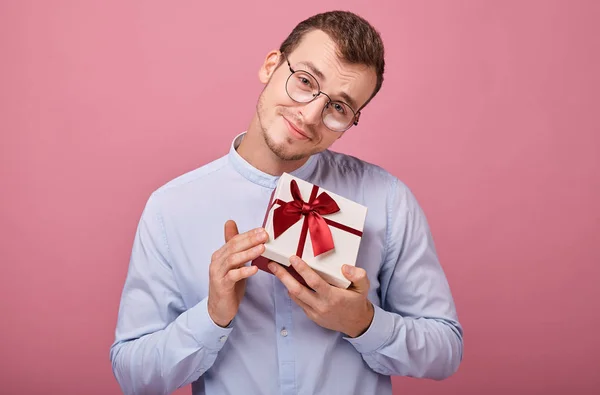 Um homem surpreso de camisa azul com óculos segura um presente em uma caixa com tampa e arco vermelho em suas mãos, olhando para o presente. Férias, regozijo. Sorrindo, felicidade. Dá-me um presente. O tipo baixou a cabeça. . — Fotografia de Stock